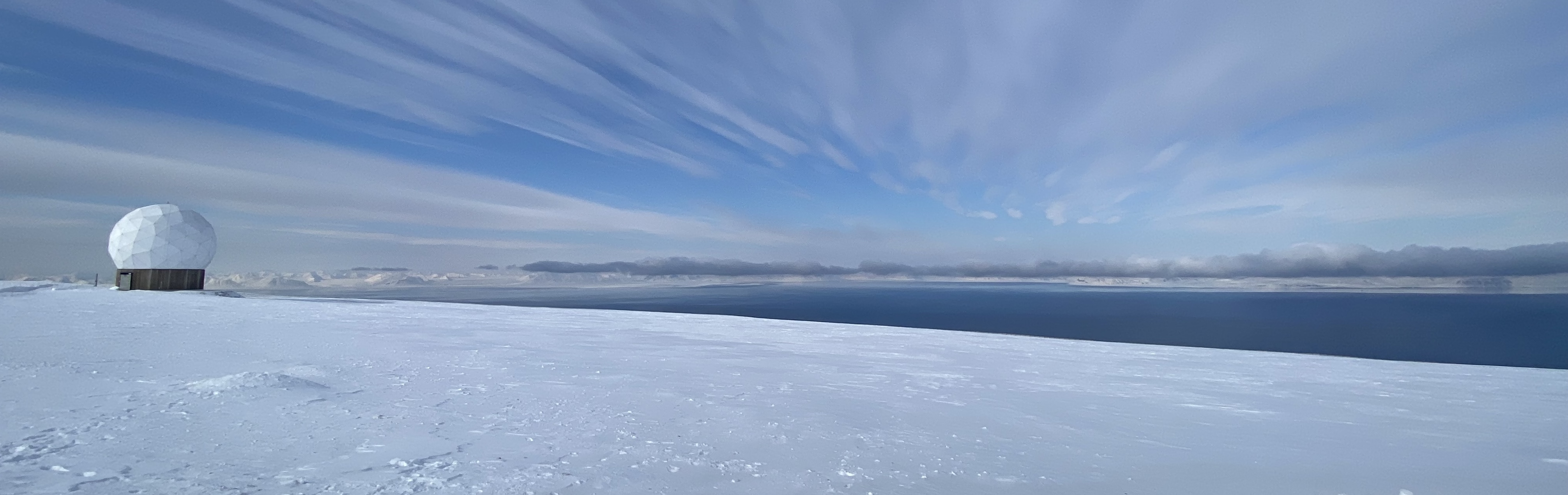 Lauren Hoffman in a fur-lined parka on a snow-covered plateau in Svalbard, with a satellite receiving station and a fjord and mountains in the background